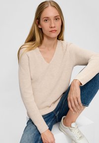 Beige geribde breiselsweater met een V-hals, vergezeld van blauwe denim jeans en witte sneakers, zittend op een wit platform.