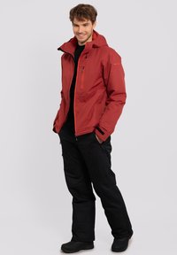 Chaqueta impermeable roja con acentos naranjas, cremallera frontal y capucha, combinada con pantalones negros y botas negras. Conjunto de ropa de exterior.