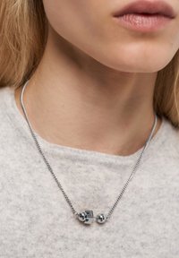 Sølv kæde med et lille geometrisk vedhæng, der har en kube og to runde perler, båret på en lysfarvet top.