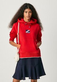 Felpa rossa con tasca frontale, cappuccio con cordino e patch logo blu e bianco. Indossata con una gonna svasata blu navy e una piccola borsa bianca.