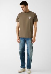 Olivgrünes T-Shirt mit kurzen Ärmeln und rundem Ausschnitt, mit einem Logo auf der Brust. Kombiniert mit ausgewaschenen blauen Jeans und weißen Sneakern.