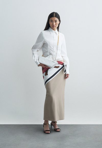 Kobieta w białej bluzce w fałdy, beżowej długiej spódnicy z zawiązaną wzorzystą chustą, czarnych sandałach na obcasie z paskami oraz białej geometrycznej torebce na łańcuszku.