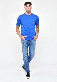 T-shirt in cotone blu con collo rotondo e maniche corte. Presenta una piccola tasca nera con un logo. Abbinata a jeans azzurri chiari e sneakers navy.