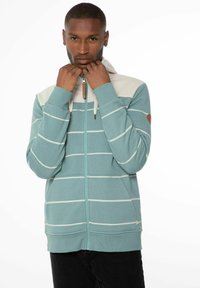 Donsjack met rits in teal met witte horizontale strepen, met een beige capuchon, ribbels aan de mouwen en een houten ritsoptrekker.