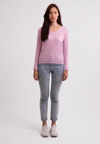 Rosa v-ringad tröja, långa ärmar, kombinerad med ljus tvättade jeans. Modellen står i neutralt ljus, iklädd vita sneakers.