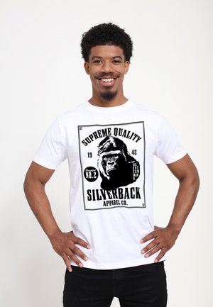 Uomo sorridente con capelli corti e ricci che indossa una maglietta bianca con una grafica nera di gorilla e il testo "Supreme Quality Silverback Apparel Co."