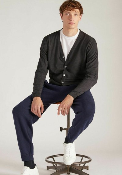 Donkergrijze cardigan met knopen, gedragen over een wit T-shirt. Gecombineerd met marineblauwe joggingbroek en witte sneakers. Model zittend op een kruk.