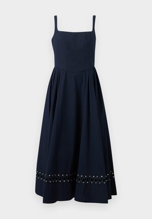 Nő fekete ujjatlan midi ruhát visel, négyzet alakú nyakkivágással, szemlyuk részletekkel az aljánál, és bézs alacsony sarokkal rendelkező cipővel, pántos hátoldallal.