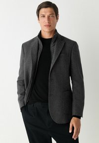 Blazer grigio a spina di pesce con revers a punta, chiusura a un bottone e due tasche anteriori, indossato sopra un maglione e pantaloni neri.