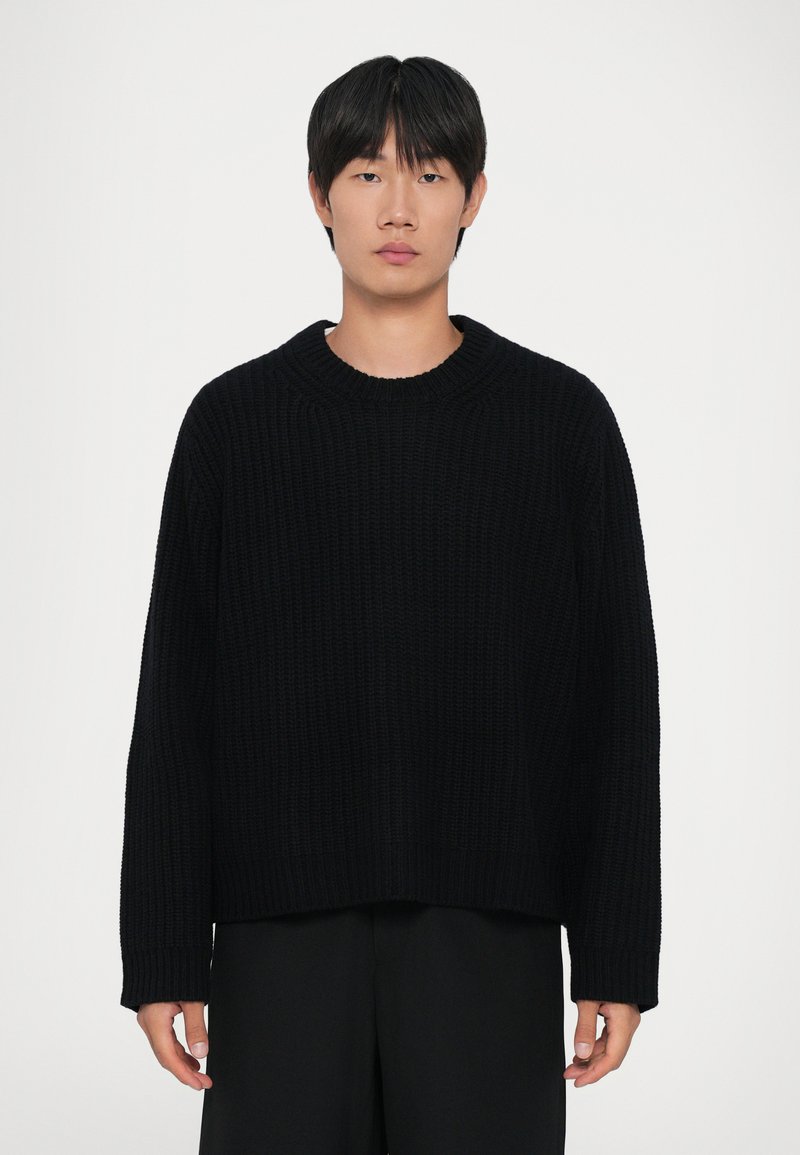 Giovane uomo con capelli neri lisci, che indossa un maglione nero lavorato a maglia largo e pantaloni neri, in piedi di fronte a uno sfondo chiaro e uniforme.