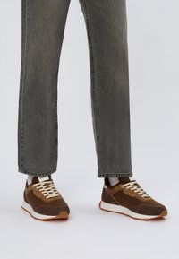 Baskets brunes avec une tige en daim et en mesh, lacets beiges et semelle en caoutchouc orange. Portées avec un jean droit gris.