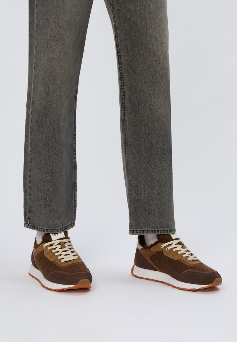 Baskets brunes avec une tige en daim et en mesh, lacets beiges et semelle en caoutchouc orange. Portées avec un jean droit gris.