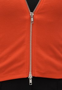 Κόκκινο ριμπ τοπ με κεντρικό ασημένιο φερμουάρ και τράβηγμα, υφασμάτινη υφή και κομψή cropped σχεδίαση. Το μαύρο φόντο αναδεικνύει το ένδυμα.