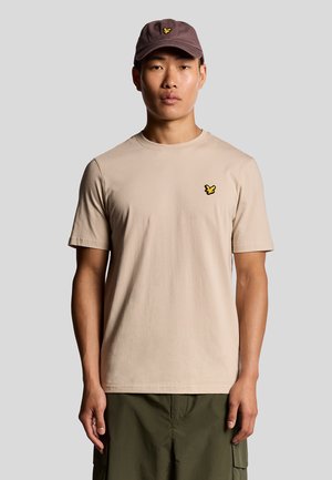 Beige cotton t-shirt with short sleeves, featuring a small black and yellow butterfly logo on the chest. Paired with olive green shorts.