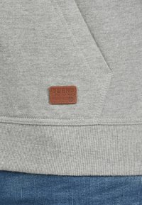 Gray sweatshirt with a ribbed hem and a leather patch labeled "BLEND." Soft fabric texture and angular design details. Denim jeans visible below.