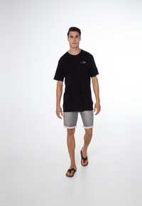 Zwarte katoenen t-shirt met een klein gekleurd logo, vervaagde grijze denim shorts en bruine flip-flops. De outfit is casual en ontspannen.