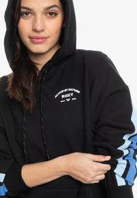 Sudadera negra con capucha, con texto en blanco "Active by Nature" y "Roxy". Rayas de acento azul en las mangas, textura suave y corte estándar.