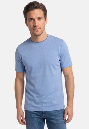 Mand med kort brunt hår iført en lyseblå t-shirt med korte ærmer og rund hals samt blå jeans mod en ensfarvet baggrund.