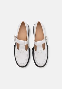 Mocassins en cuir blanc avec lanières en T, semelles noires et fermetures à boucle, arborant des logos embossés sur des semelles intérieures lisses de couleur tan.