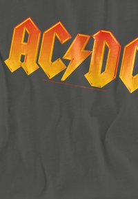 Dunkelgraues T-Shirt mit auffälligem gelb-orangem Schriftzug "AC/DC" und einem Grafikelement in Form eines Blitzes. Der Text hat einen leicht abgenutzten Look.