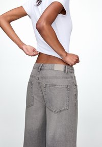 Pantalons larges en denim gris avec un patch à la taille, dotés de deux poches arrière et d'une coupe ample, associés à un t-shirt blanc ajusté.