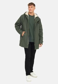 Mladý muž v zelené parce s kožíškem pod kapucí, oblečený v tmavě zeleném svetru, černých kalhotách a bílých teniskách, stojí sebevědomě.