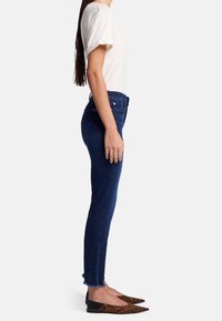 Mujer vistiendo una blusa blanca de manga corta, jeans ajustados azul oscuro con dobladillos deshilachados, y zapatos planos de punta con estampado de leopardo, de pie de lado sobre un fondo blanco.
