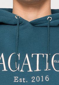 Felpa teal realizzata in morbido tessuto, con cappuccio regolabile e grande scritta ricamata in rosa chiaro che dice "VACATION Est. 2016."