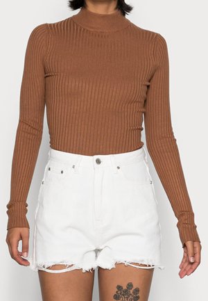 Personne portant un haut marron à manches longues côtelé, rentré dans un short en denim blanc taille haute avec des ourlets effilochés, et un tatouage floral sur la cuisse.