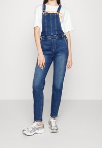 Jeanslatzhosen mit verstellbaren Trägern, schlanker Passform und einem verblassten blauen Farbton. Getragen über einem weißen T-Shirt und kombiniert mit grauen Sneakers.