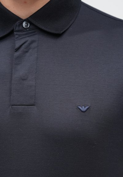 Tumši zils polo krekls ar teksturētu, kontrastējošu apkakli, centrālu pogu joslu un nelielu izšūtu logotipu krūtīs.