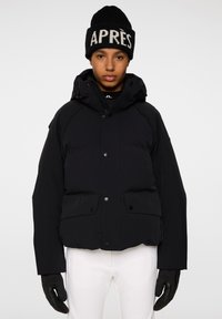 Svart vadderad jacka med huva, tryckknappar och två framfickor. Bärs med en svart stickad mössa med vit "APRÈS" text.