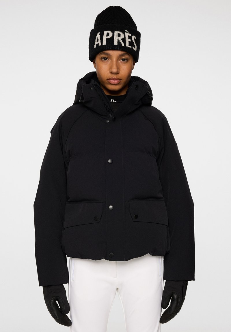 Svart vadderad jacka med huva, tryckknappar och två framfickor. Bärs med en svart stickad mössa med vit "APRÈS" text.
