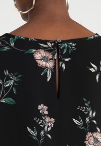 Top negro con patrón floral en rosa y verde, que presenta un detalle en forma de ojal en la parte posterior, cierre de botón y una textura de tela suave.