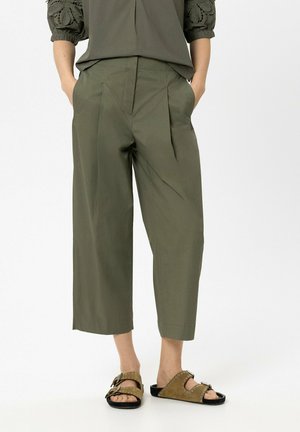 Person trägt olivgrüne, gekürzte Hosen mit weitem Bein und steckt die Hände in die Taschen, dazu braune Pantoletten mit zwei Schnallenriemen.
