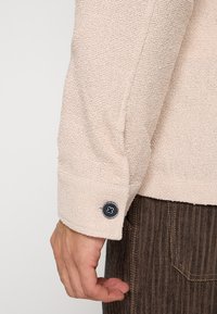 Beige langarmshirt met textuur en een zwarte knoop op de manchet, gecombineerd met donderbruine geribbelde broek.