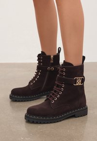 Mujer que lleva botas de ante marrón oscuro, con cordones y cremalleras laterales, además de una hebilla dorada decorativa, sobre un suelo liso.