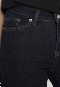 Mörkblå denimjeans med orange sömmar. Har en framficka och metallknappstängning. Materialet är slätt med en lätt glans.