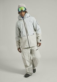 Light grey snow jacket with a zippered front, two zip pockets, and a hood. Paired with matching light grey snow pants and black boots.