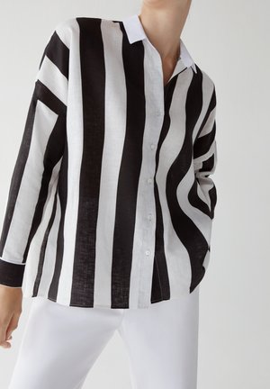 Osoba ubrana w czarno-białą koszulę w pionowe paski na guziki z długimi rękawami oraz białe spodnie na jednolitym tle.