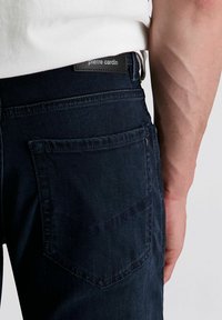 Nahaufnahme der Hüfte einer Person, die dunkelblaue Jeans mit einer Gesäßtasche und einem sichtbaren "Pierre Cardin"-Label sowie ein weißes Hemd trägt.
