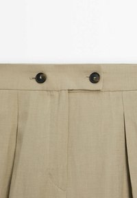 Ceinture d'un pantalon beige avec deux boutons foncés et des plis en dessous, mettant en valeur le détail de la fermeture boutonnée.