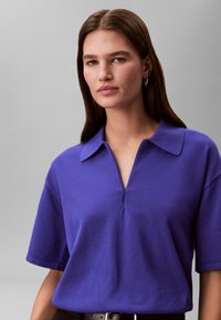 Γυναίκα με καστανά ίσια μαλλιά, που φοράει ασημένια κρίκους σκουλαρίκια και μωβ πουκάμισο με κοντά μανίκια και γιακά, μπροστά σε γκρίζο φόντο.