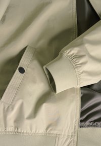 Chaqueta bomber de color caqui claro, fabricada en un tejido suave y ligero. Cuenta con puños acanalados y cierre de cremallera, además de un detalle de bolsillo con snap lateral.