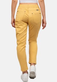 Pantalon stretch jaune avec une taille élastique, deux poches arrière et des détails cousus, associé à des baskets blanches.