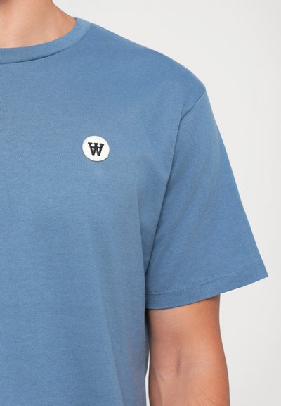 Blaues Baumwoll-T-Shirt mit Rundhalsausschnitt und kurzen Ärmeln, ausgestattet mit einem weißen gestickten runden Patch mit dem schwarzen Buchstaben "W."