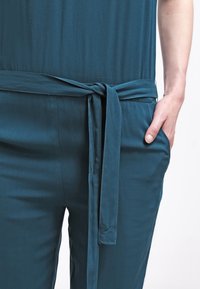Pantalons teal en tissu lisse avec une taille nouée. Comprend deux poches latérales et une coupe décontractée, mettant en avant un design simple.