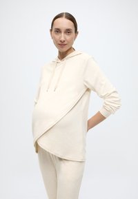 Sweat à capuche de maternité beige clair avec un design portefeuille, manches longues, cordon de serrage ajustable et texture douce. Associé à un pantalon assorti.