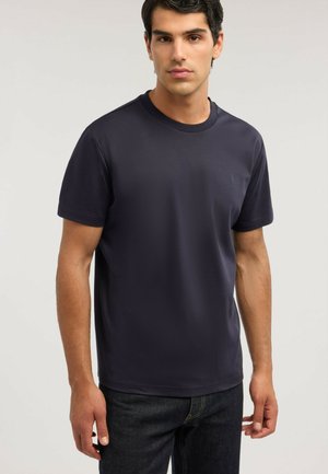 Młody mężczyzna ubrany w gładki ciemnoniebieski krótki rękaw t-shirt i ciemne dżinsy, stojący na jasnoszarym tle.