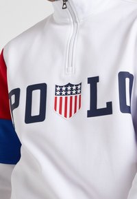 Bílá mikina se čtvrtinovým zipem s červenými a modrými akcenty. Na přední straně je velké logo "POLO" a štít s hvězdami a pruhy uprostřed.
